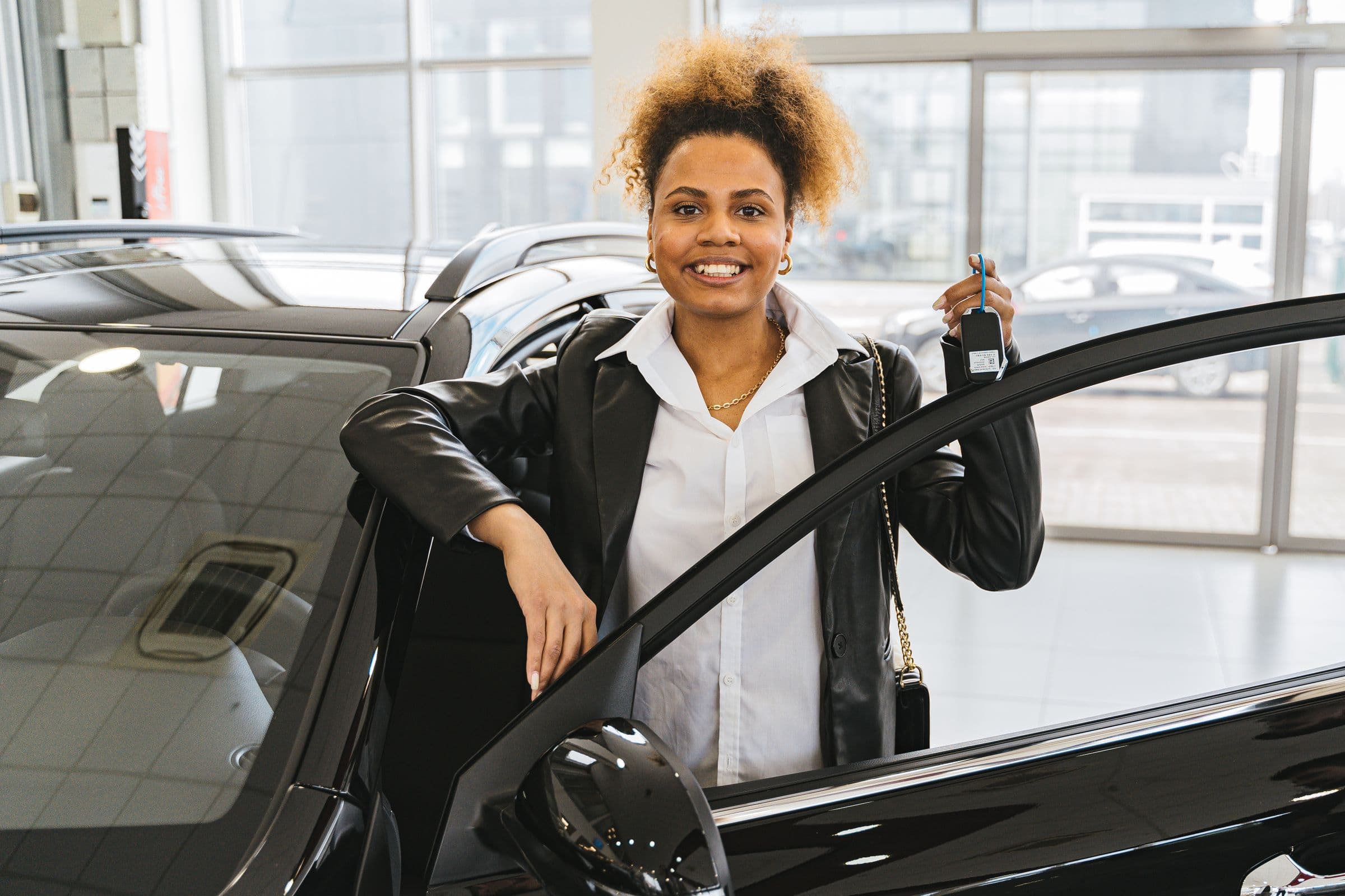 A smiling woman holds up the keys to her car at a dealership showroom.